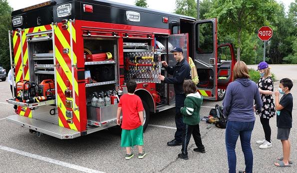 Children in summer play clothes and a woman in jeans watch as a Richfield firefighter explains equipment on a fire truck. 